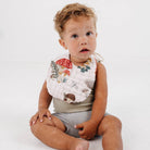 A toddler sitting on a white studio floor wearing a white bib with a colorful forest print of mushrooms, leaves, and a squirrel, paired with a gray ribbed tank top and shorts.