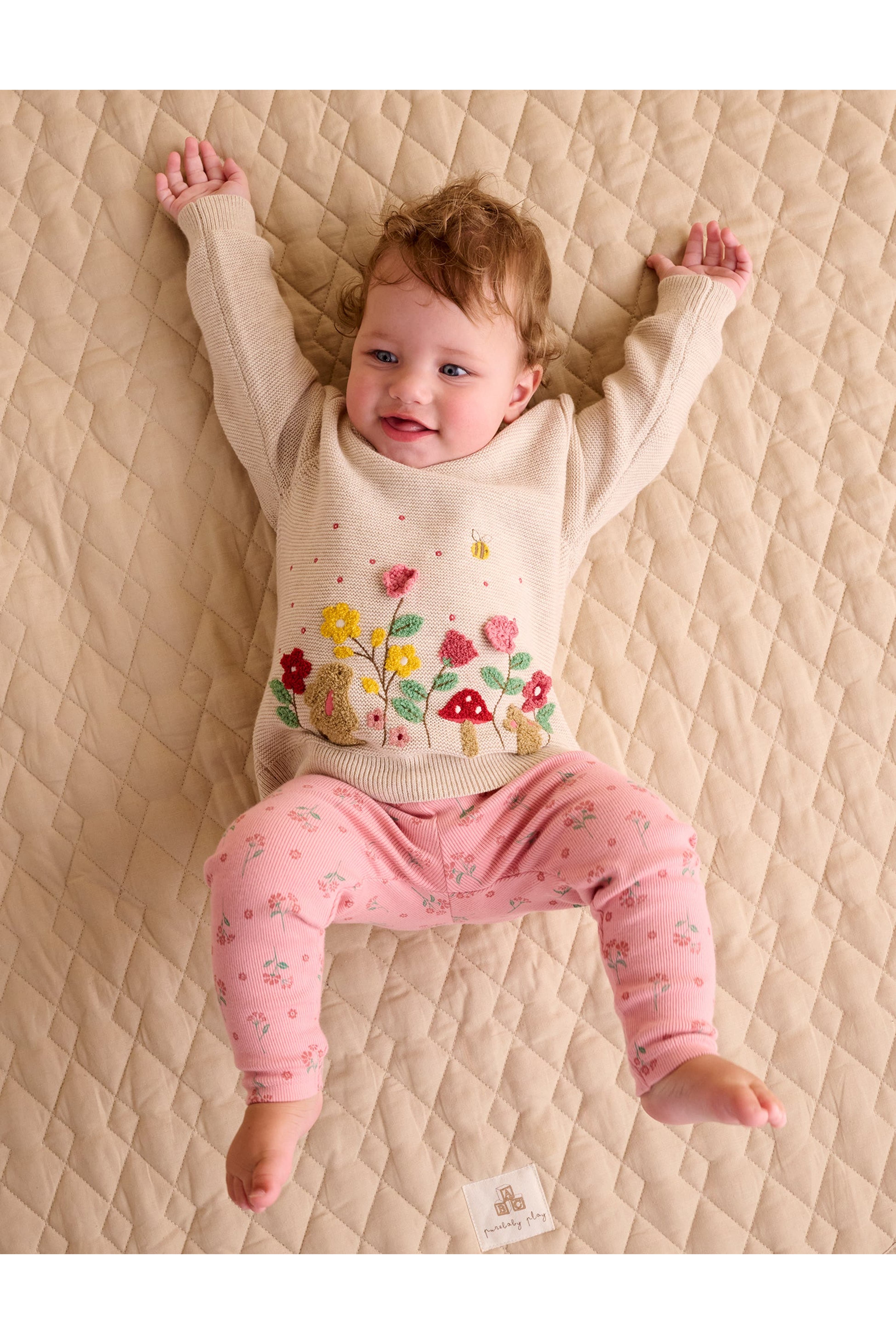 A baby lying on a quilted beige mattress, wearing a cream knit sweater with a colorful mushroom and flower design and pink patterned leggings.