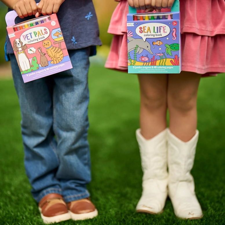 Two children standing on green grass holding coloring book boxes. The child on the left wears blue jeans and brown shoes and holds a 'Pet Pals Coloring Book'.