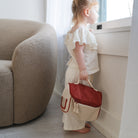 Child holding a red and beige bag in a room with a couch and window.