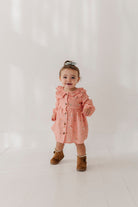 A toddler girl wearing a dusty pink long-sleeve button-up dress with a ruffled collar and small floral print, paired with brown boots, standing against a bright white studio background.