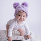 Baby wearing a purple knit hat with two large pom-poms, sitting on a white fluffy surface.