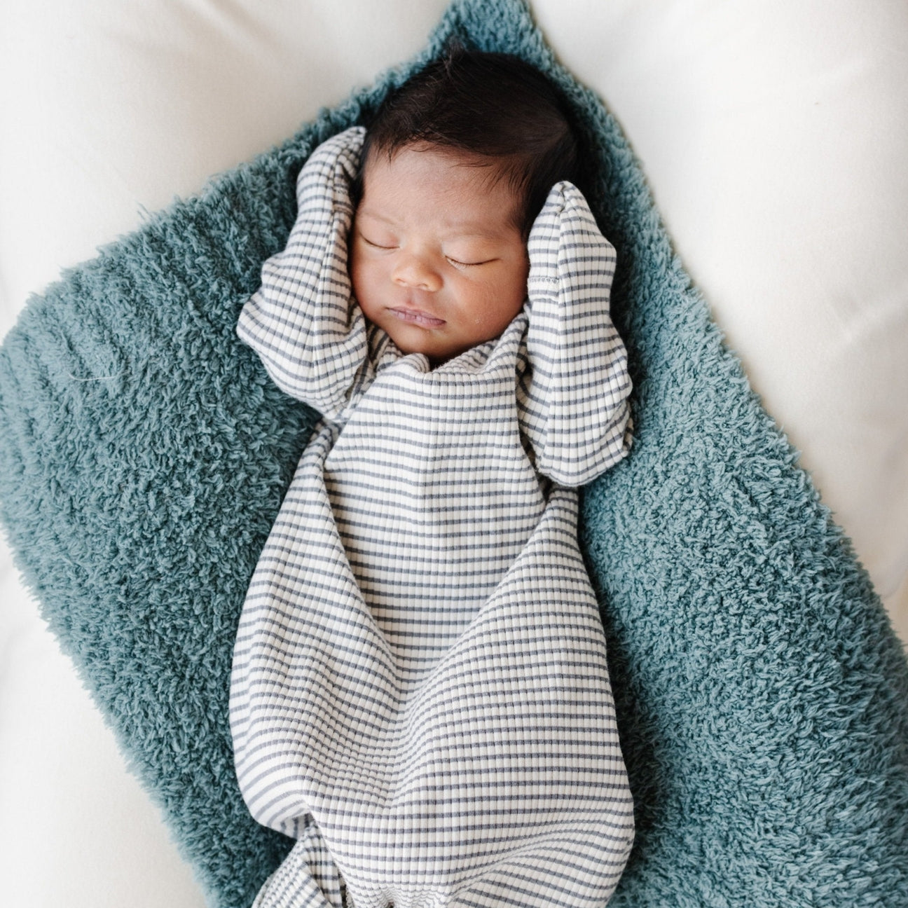 A sleeping newborn baby wrapped in a soft, striped muslin swaddle blanket, nestled on a plush teal blanket over white bedding.