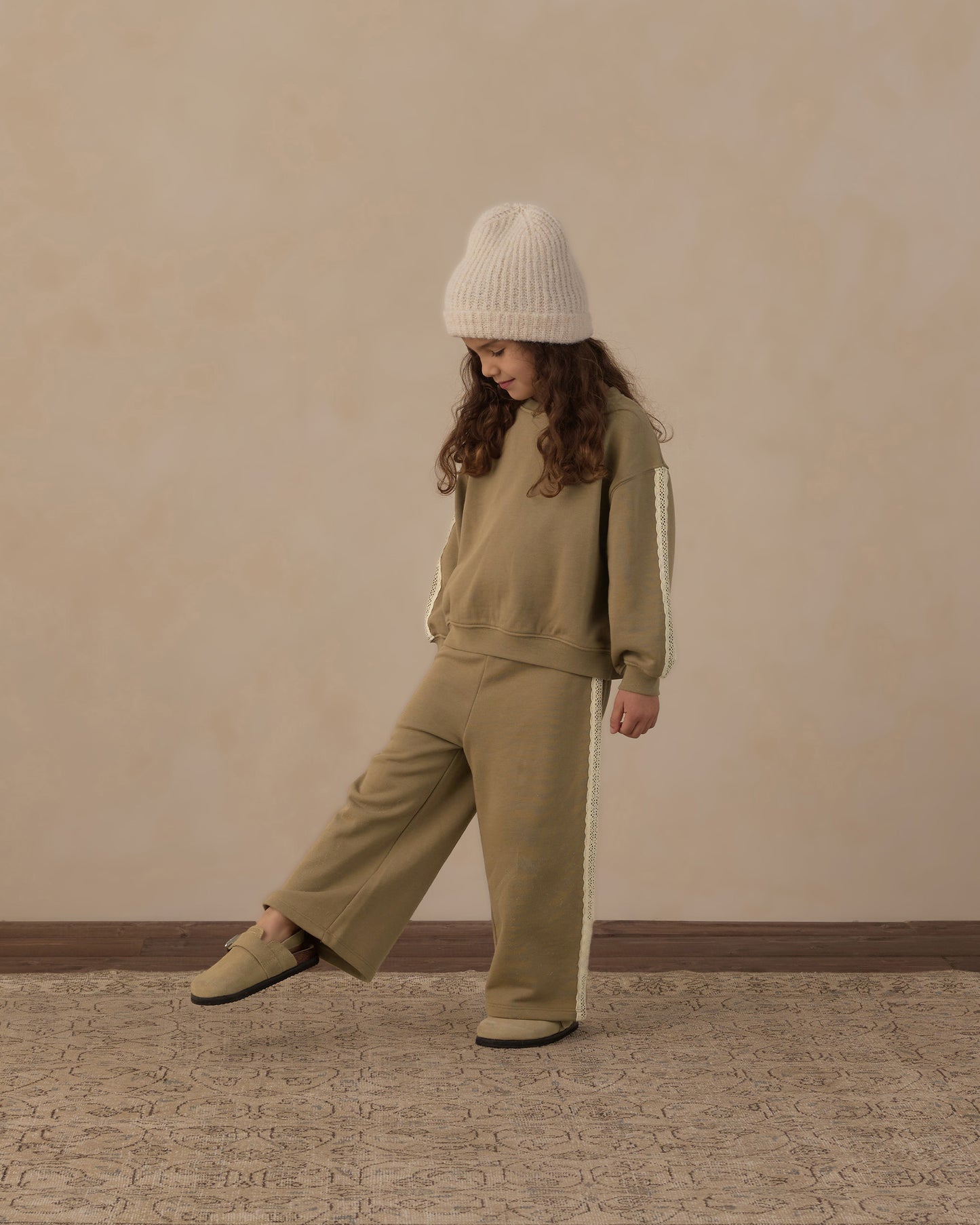 A child wearing a matching beige sweatshirt and wide-leg pants set with white side stripes, a cream knit beanie, and beige shoes, standing against a textured beige wall.