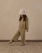 A child wearing a matching beige sweatshirt and wide-leg pants set with white side stripes, a cream knit beanie, and beige shoes, standing against a textured beige wall.