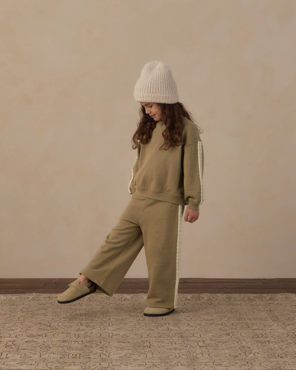 A child wearing a matching beige sweatshirt and wide-leg pants set with white side stripes, a cream knit beanie, and beige shoes, standing against a textured beige wall.