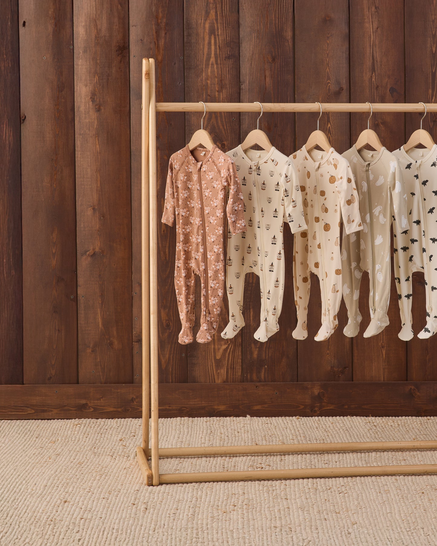 Five baby footed onesies in soft neutral tones hang on a wooden clothing rack against a dark wood plank wall. The onesies include pink floral, cream tree prints, and cream polka dot patterns.
