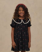 A young girl with curly brown hair wearing a black short-sleeve dress with a white lace Peter Pan collar and a red floral print, paired with a red headband, standing against a textured beige background.
