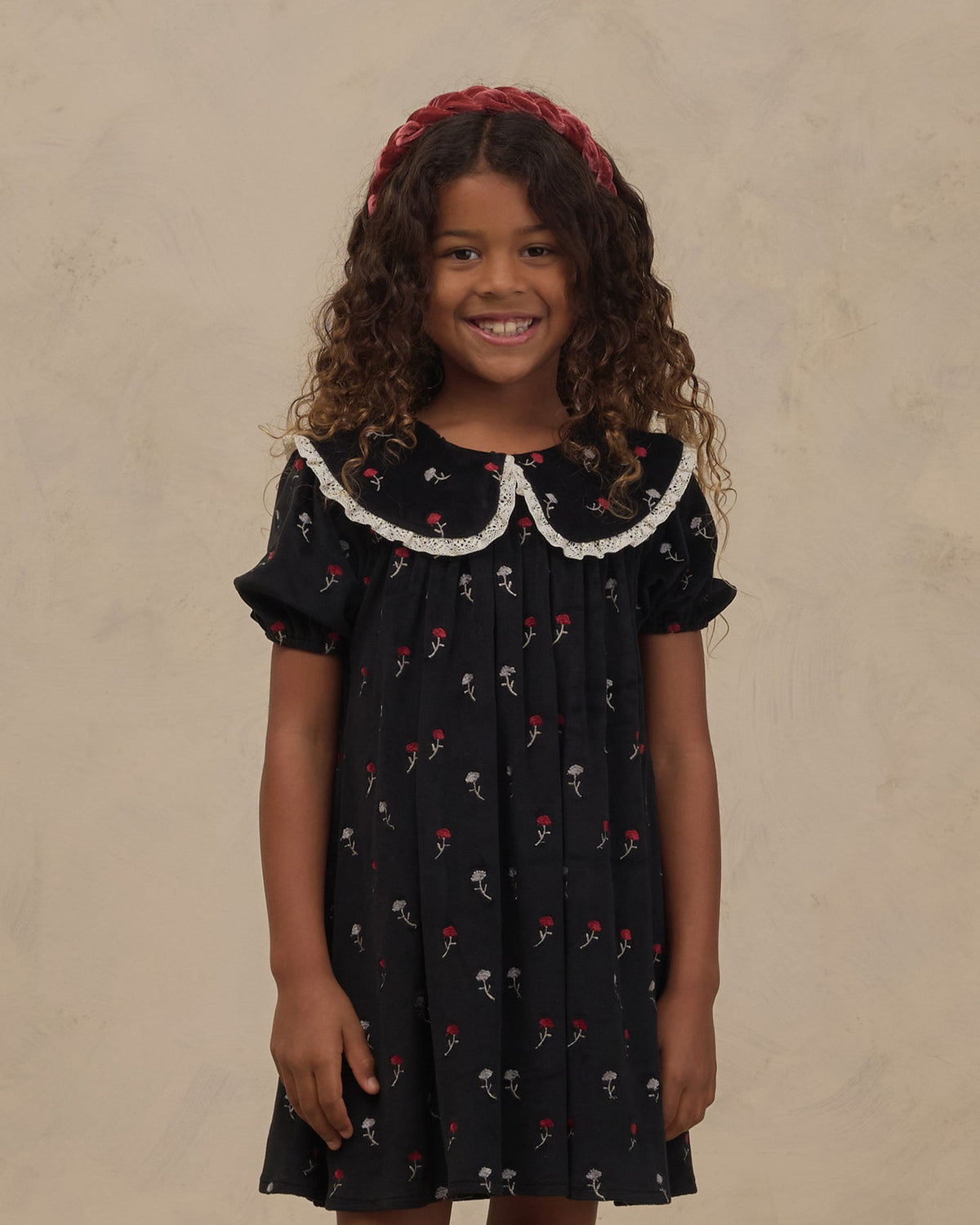 A young girl with curly brown hair wearing a black short-sleeve dress with a white lace Peter Pan collar and a red floral print, paired with a red headband, standing against a textured beige background.