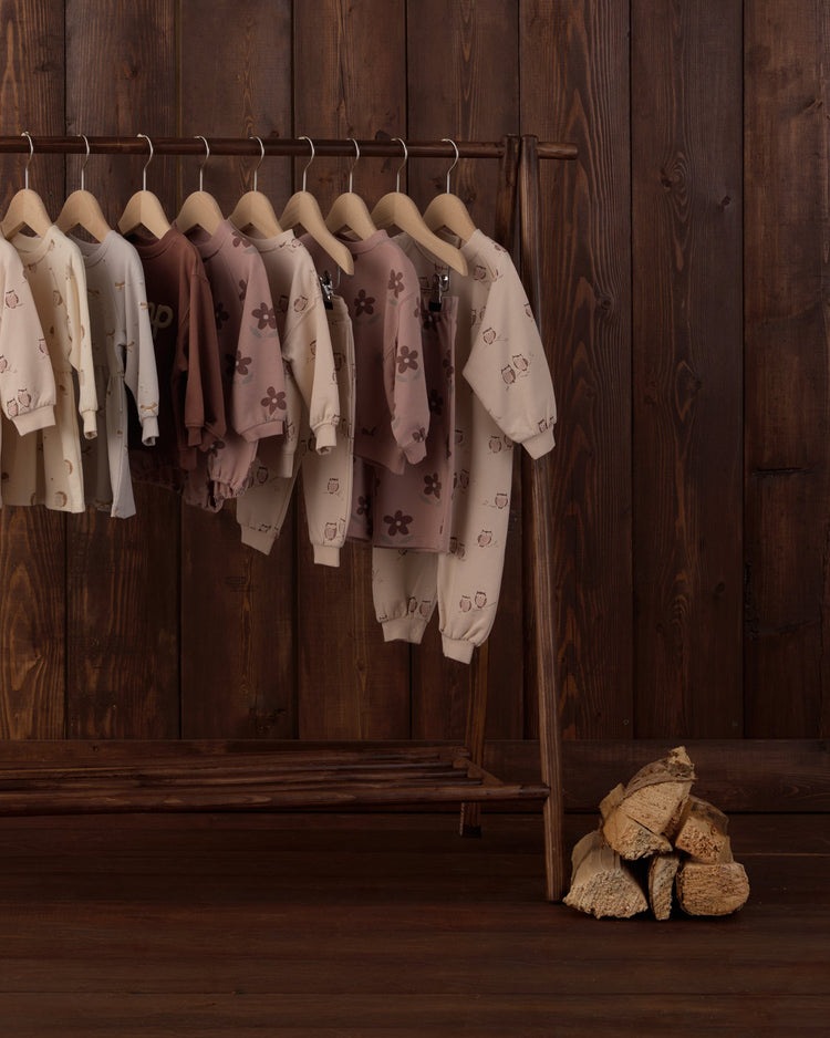 Rylee + Cru neutral-toned children's clothing collection featuring cozy sweatshirts, track pants, and jumpsuits with subtle floral prints displayed on a wooden clothing rack.