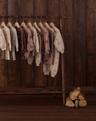 Rylee + Cru neutral-toned children's clothing collection featuring cozy sweatshirts, track pants, and jumpsuits with subtle floral prints displayed on a wooden clothing rack.