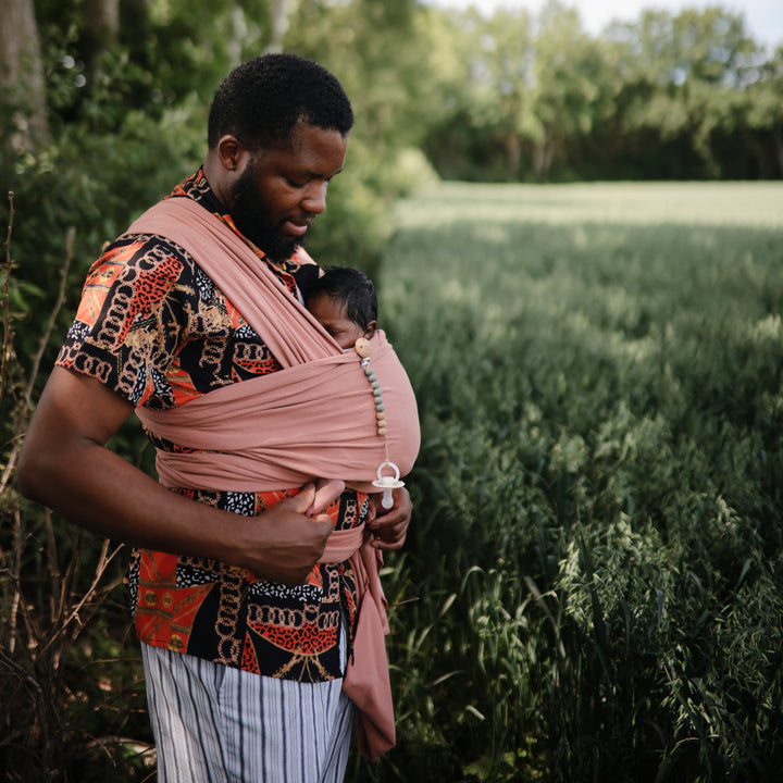 Man holding a baby in a sling outdoors with greenery in the background
