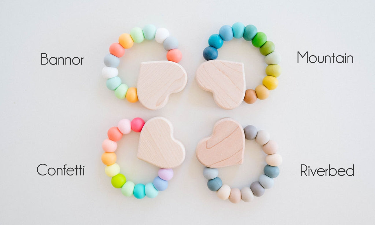 Four wooden teething rings arranged in a 2x2 grid on a white background, each with pastel-colored beads and a light wood heart-shaped teether. Text labels identify the variants as Bannor, Mountain, Confetti, and Riverbed.