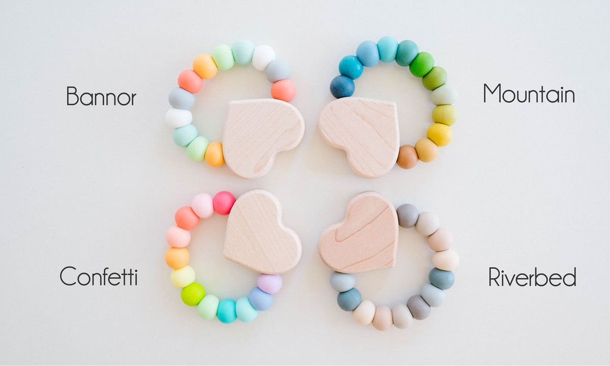 Four wooden teething rings arranged in a 2x2 grid on a white background, each with pastel-colored beads and a light wood heart-shaped teether. Text labels identify the variants as Bannor, Mountain, Confetti, and Riverbed.