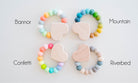 Four wooden teething rings arranged in a 2x2 grid on a white background, each with pastel-colored beads and a light wood heart-shaped teether. Text labels identify the variants as Bannor, Mountain, Confetti, and Riverbed.