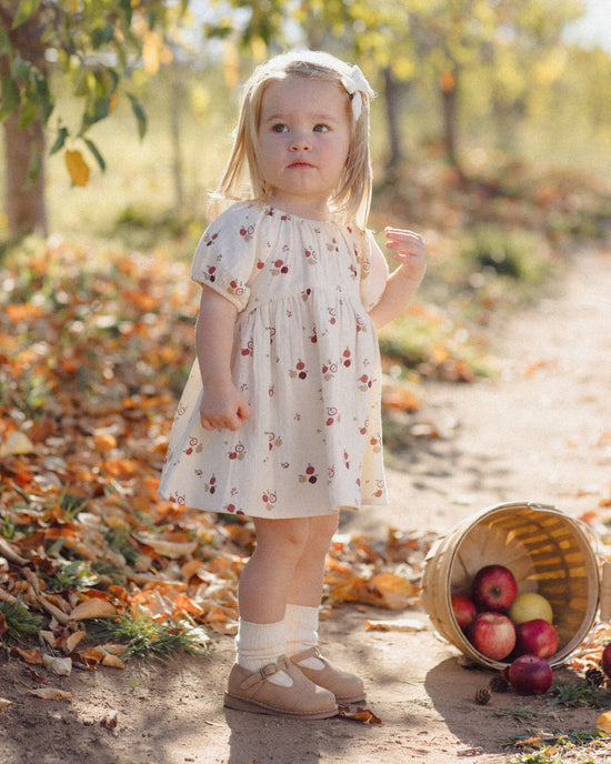 A toddler girl stands on a dirt path outdoors wearing a cream short-sleeve dress with a red cherry print. A tipped basket of red and yellow apples sits on the path beside her, with fallen leaves and trees in the background.