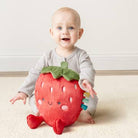 Baby holding a strawberry-shaped plush toy on a light-colored floor.