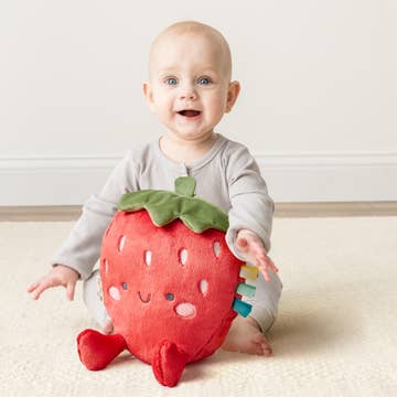 Baby holding a strawberry-shaped plush toy on a light-colored floor.