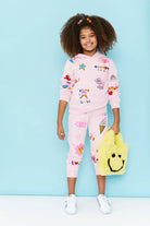 A young girl with curly hair stands against a light blue studio background wearing a pink hooded sweatshirt and matching jogger pants covered in colorful cartoon patches. She is holding a yellow fuzzy smiley-face bag.