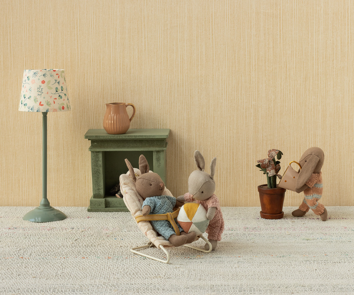 Two toy Maileg micro plush rabbits in a nursery scene, with one rabbit sitting in an off-white bouncy chair holding a small ball while another rabbit stands nearby.
