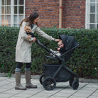 Woman with a baby in a stroller and a dog, standing on a sidewalk with a brick building in the background.