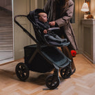 Black stroller with a baby inside, being pushed by a person in a home setting.
