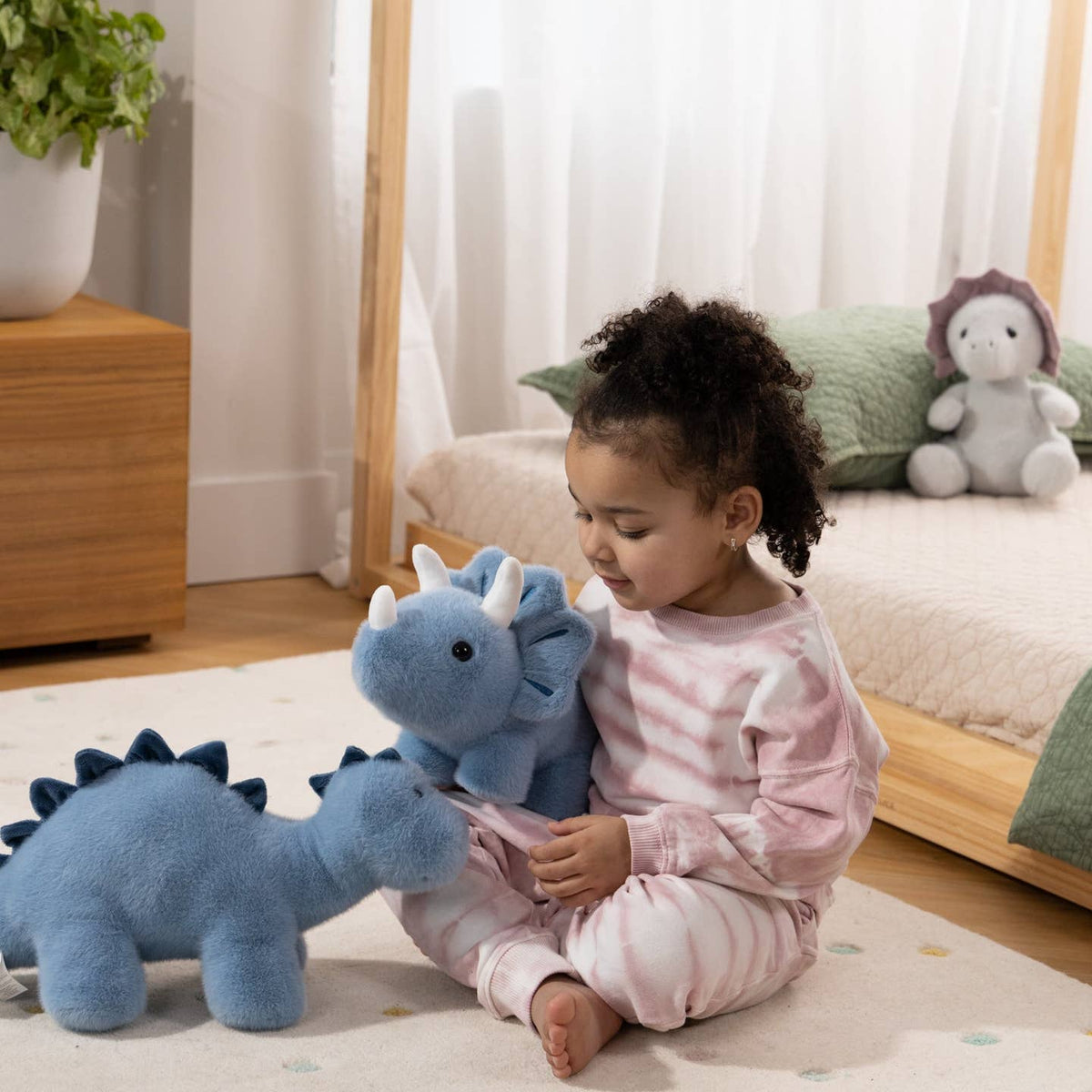 A young child wearing pink and white striped pajamas sits on a carpeted floor, holding a blue plush dinosaur toy next to another blue dinosaur plush in a bright bedroom setting.