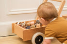 A baby in a mustard yellow ribbed romper crawls beside a wooden toy cart filled with plush stuffed animals on a light wood floor.
