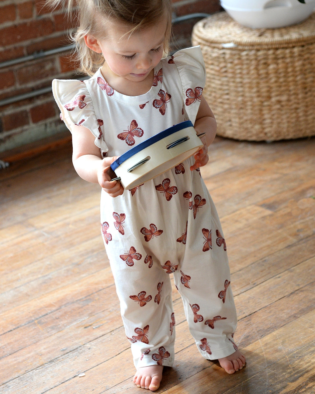 Child wearing a white romper with butterfly patterns, holding a toy on a wooden floor.