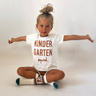 A young girl with her hair in a high bun sits cross-legged with arms outstretched, wearing a white t-shirt that reads 'KINDER GARTEN squad' in brown lettering, brown shorts, and white knee-high socks with brown and teal accents.