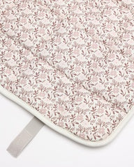 A close-up of a quilted baby blanket or mat featuring a delicate pink and brown floral pattern on a light cream background, finished with a white border and a gray strap.