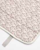 A close-up of a quilted baby blanket or mat featuring a delicate pink and brown floral pattern on a light cream background, finished with a white border and a gray strap.