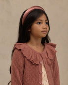 A young girl with long dark hair wearing a dusty rose pink ribbed cardigan with a ruffled collar and matching buttons, along with a matching pink satin headband, set against a neutral textured beige background.