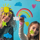 Two children smiling and painting on a blue glass window with colorful crayon drawings of a rainbow, sun, clouds, and hearts.