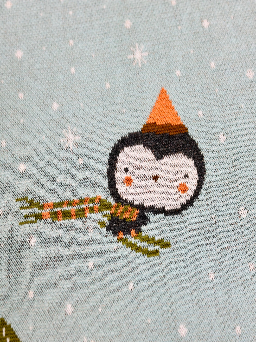 Cross-stitched penguin holding a bundle of sticks on a light blue background with snowflakes.