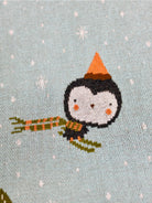 Cross-stitched penguin holding a bundle of sticks on a light blue background with snowflakes.