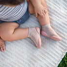 A close-up lifestyle shot of a baby sitting on a textured cream knit blanket outdoors, wearing light pink knit socks with solid pink toes.