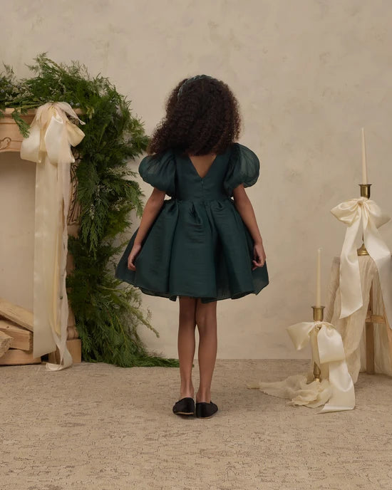 A young girl with curly dark hair is shown from behind wearing a dark teal green puff-sleeve dress. She is standing in a styled indoor setting with a textured beige wall, a greenery and cream ribbon fireplace on the left, and matching cream ribbon candlesticks on the right.