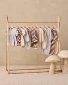 A wooden baby clothing rack holding multiple baby garments in soft neutral tones, set against a warm beige wall with a textured rug and small mushroom-shaped side tables.