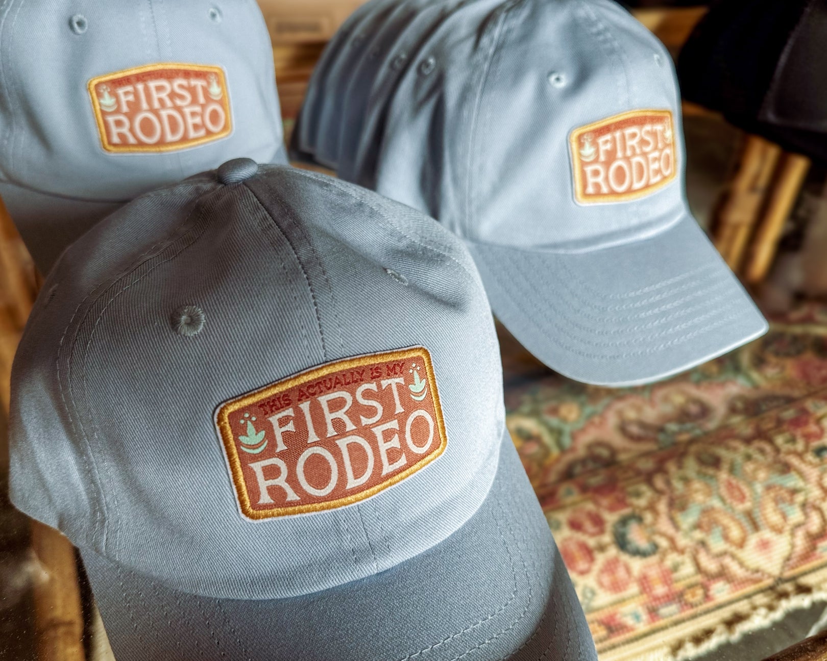 Two gray baseball caps with 'First Rodeo' logo on a patterned surface