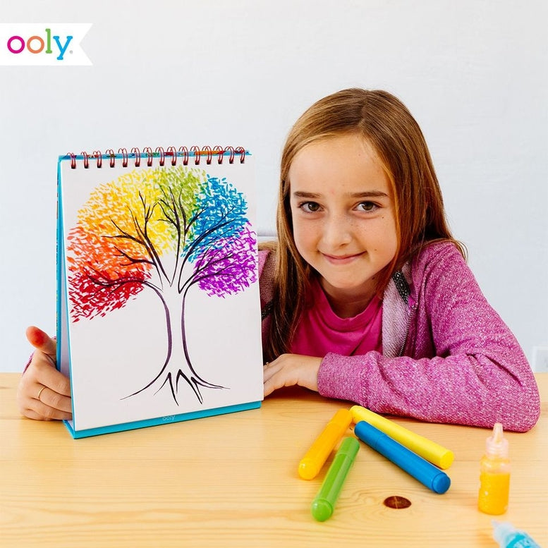 A young girl with long light brown hair smiles while holding a colorful tree drawing in an Oooly. spiral-bound sketchbook.