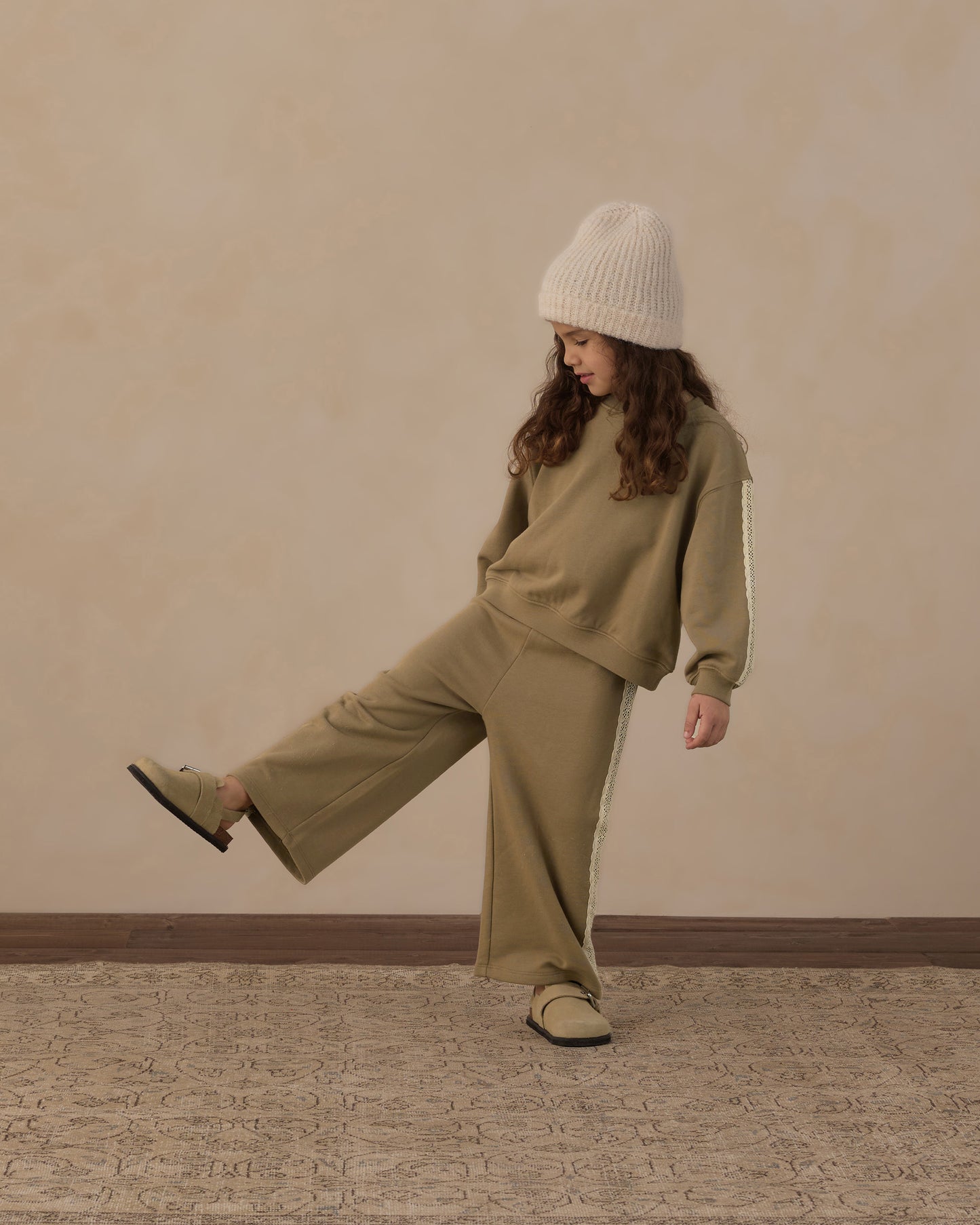A child wearing a matching khaki green sweatshirt and wide-leg sweatpants set with white side stripes, a cream knit beanie, and beige slip-on shoes, posing against a textured beige wall.