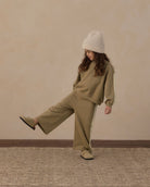 A child wearing a matching khaki green sweatshirt and wide-leg sweatpants set with white side stripes, a cream knit beanie, and beige slip-on shoes, posing against a textured beige wall.