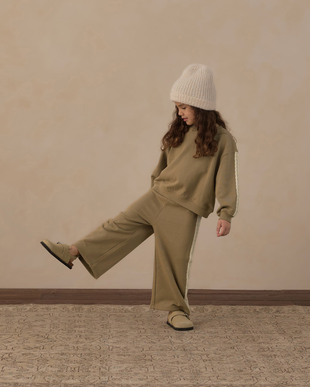 A child wearing a matching khaki green sweatshirt and wide-leg sweatpants set with white side stripes, a cream knit beanie, and beige slip-on shoes, posing against a textured beige wall.