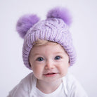 Child wearing a purple knit hat with two large pom-poms on a light background