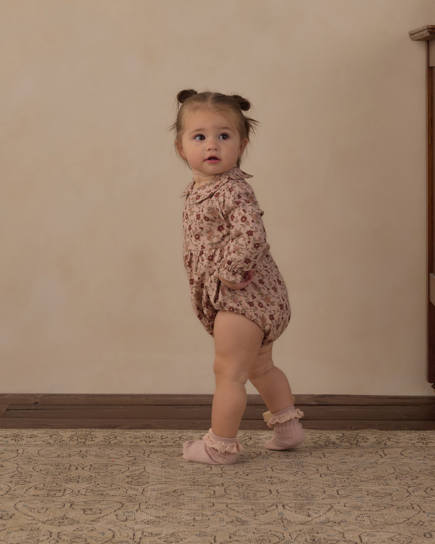 A toddler girl wearing a dusty pink floral long-sleeve romper and matching pink booties, standing on a patterned rug against a beige wall.