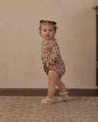 A toddler girl wearing a dusty pink floral long-sleeve romper and matching pink booties, standing on a patterned rug against a beige wall.