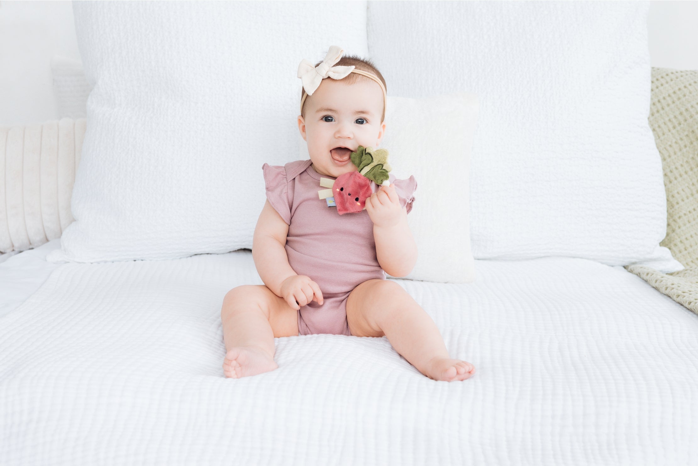 Baby in a pink outfit sitting on a white bed holding a toy.