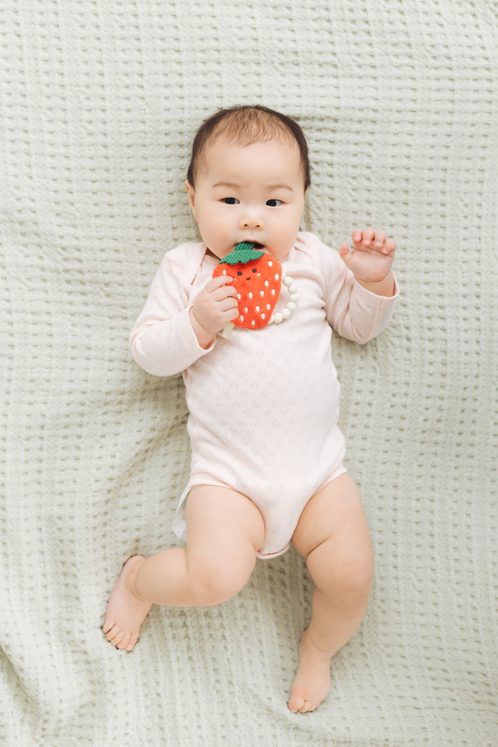 Baby in a white onesie holding a strawberry-shaped toy on a light green textured blanket