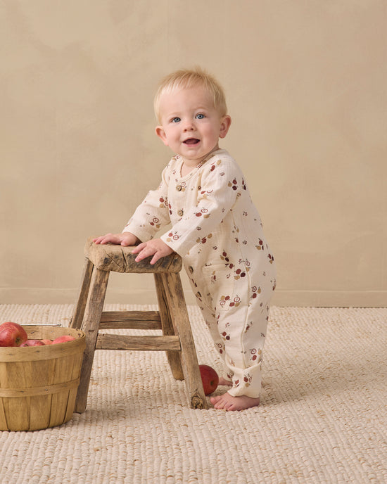 A baby standing on a woven rug next to a small wooden stool, wearing a cream-colored long-sleeve footless onesie with a brown floral pattern. A wooden basket of red apples sits on the floor to the left.